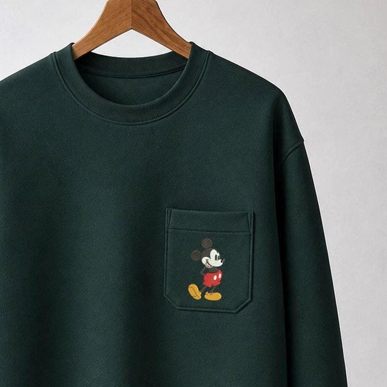 May include: A dark forest green crewneck sweatshirt featuring a single chest pocket. The pocket displays a small, embroidered graphic of Mickey Mouse, depicted with his classic black ears, white face, red shorts, and yellow shoes.