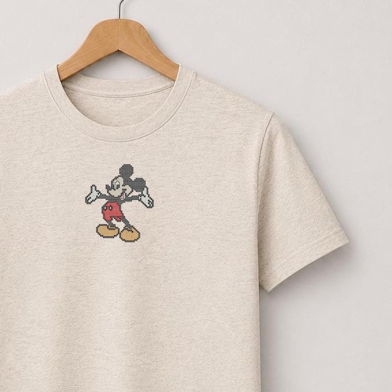 Pu&ograve; includere: Una t-shirt a maniche corte color beige chiaro m&eacute;lange con un piccolo ricamo a punto croce di Topolino. Il personaggio &egrave; raffigurato in nero, bianco, rosso e giallo, con le braccia tese in una posa classica sul petto.