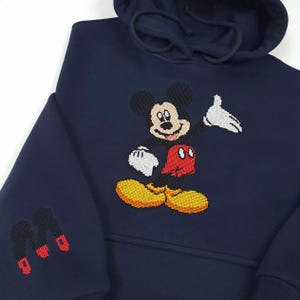 以下が含まれることがあります： 前面にクロスステッチのミッキーマウスのデザインが施されたネイビーブルーのフード付きスウェットシャツ。象徴的なキャラクターは、黒、白、赤、黄色のディテールで描かれています。左袖には、黒と赤のクロスステッチの「M」のデザインがあります。