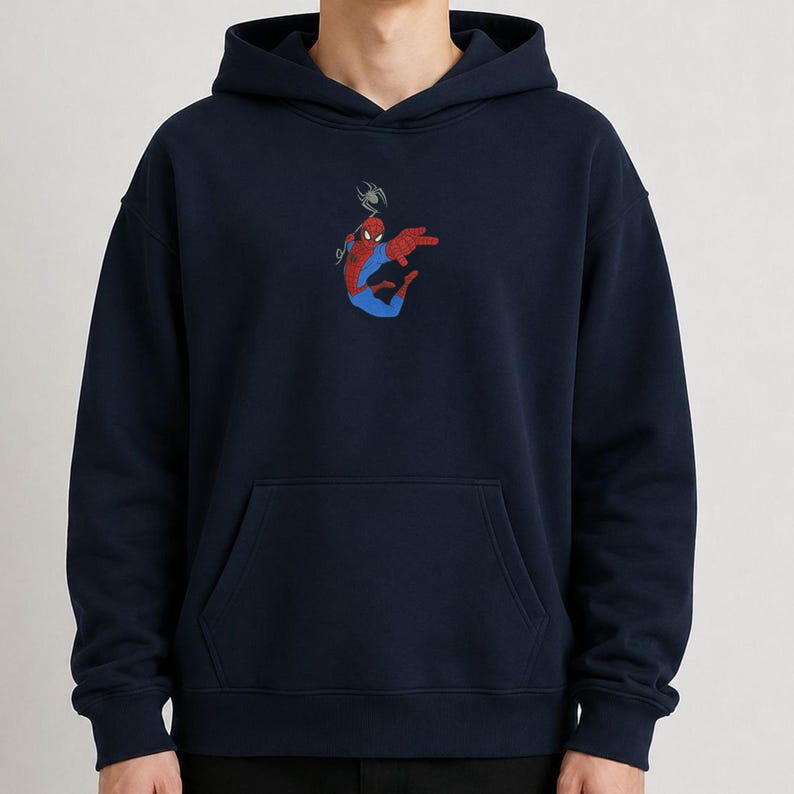 May include: A navy blue hooded sweatshirt featuring a small, centered graphic of a red and blue superhero character in a dynamic pose, suspended by a thin gray web line. The garment includes a front kangaroo pocket and long sleeves with ribbed cuffs.