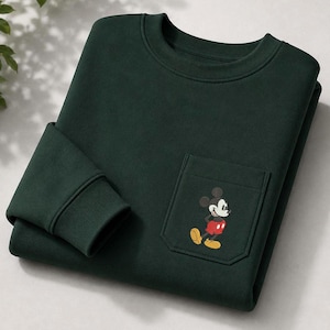 May include: A dark forest green crewneck sweatshirt featuring a single chest pocket. The pocket is adorned with a small, embroidered graphic of Mickey Mouse, depicted in his classic black, white, red, and yellow color scheme, standing upright.