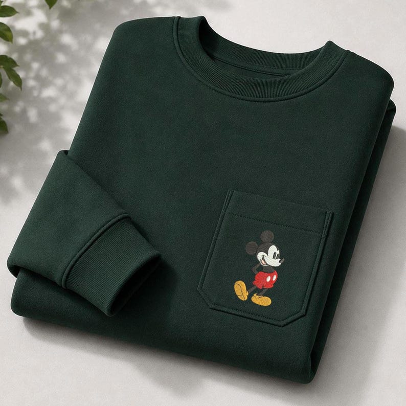 May include: A dark forest green crewneck sweatshirt featuring a single chest pocket. The pocket is adorned with a small, embroidered graphic of Mickey Mouse, depicted in his classic black, white, red, and yellow color scheme, standing upright.