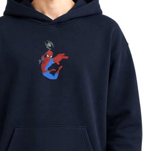 May include: A navy blue hooded sweatshirt featuring an embroidered graphic of the character Spider-Man in a red and blue suit, positioned in a dynamic pose with a small grey spider hanging from a web line above the character's head.