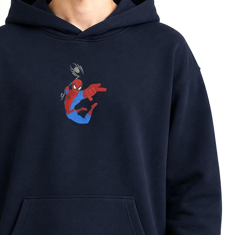 May include: A navy blue hooded sweatshirt featuring an embroidered graphic of the character Spider-Man in a red and blue suit, positioned in a dynamic pose with a small grey spider hanging from a web line above the character's head.