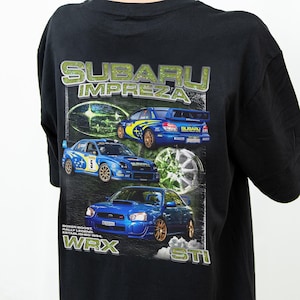 Op de afbeelding: Een zwart t-shirt met een afbeelding op de achterkant met de tekst SUBARU IMPREZA, WRX en STI. Het ontwerp bevat drie blauwe Subaru rallyauto's, een zilveren wiel en de tekst BOXER BOOST, RALLY LEGEND, ESTABLISHED 1994 in witte letters.