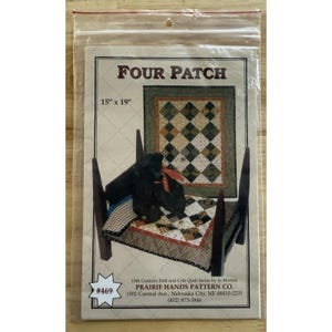 Könnte beinhalten: Eine transparente Plastikverpackung mit einem Quiltmuster. Das Muster trägt den Titel "FOUR PATCH" und misst 38 cm x 48 cm. Das Bild auf dem Muster zeigt einen kleinen Quilt auf einem Miniaturbett mit einem schwarzen Hund. Das Muster stammt von Prairie Hands Pattern Co.