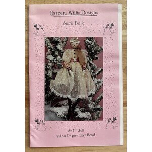 May include: A pink booklet titled "Barbara Willis Designs" featuring a doll named "Snow Belle." The doll is wearing a cream-colored dress with a lace overlay. The text reads "An 11" doll with a Paper Clay Head."