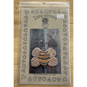 Op de afbeelding: Een knutselpatroon voor een bijenwafelpop, verpakt in een doorzichtige plastic hoes. Het patroon toont een gewatteerd bijenontwerp met bruine en gele strepen en de tekst "Love as sweet as honey bee". De verpakking is gelabeld "Dizzy & Frizzy".