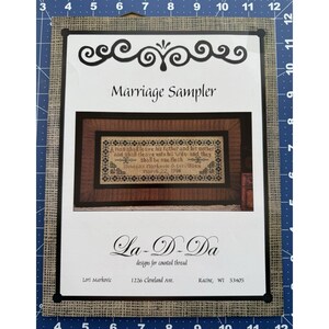 Könnte beinhalten: Ein gerahmtes Kreuzstichmuster mit dem Titel "Marriage Sampler". Das Design zeigt ein Zitat und dekorative Elemente in schwarzem Faden auf cremefarbenem Hintergrund, eingebettet in einen Rahmen in dunkler Holzoptik. Der Text "La-D-Da designs for counted thread" ist ebenfalls sichtbar.