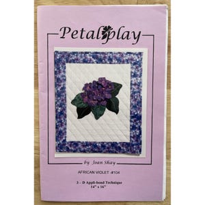 Op de afbeelding: Een lavendelkleurig patroon met een gewatteerd Afrikaans viooltje ontwerp. De afbeelding bevat de tekst "-Petal play-" en "by Joan Shay". De afmetingen zijn 35,56 cm x 40,64 cm. Het ontwerp is een 3-D Appli-bond techniek.