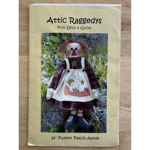 Könnte beinhalten: Eine 48 cm große Stoffpuppe namens "Punkin Patch Annie" ist auf einem beigefarbenen Papier abgebildet. Die Puppe hat ein kariertes Kleid, eine Schürze mit Kürbissen und rote Garnhaare. Das Papier trägt die Aufschrift "Attic Raggedys, Prim Dolls + Quilts."