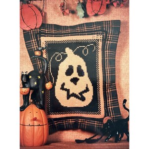 May include: A square decorative pillow featuring a central cross-stitch design of a cream-colored jack-o'-lantern on a black background, framed by a black and orange plaid fabric border. The pillow is surrounded by orange and black pumpkin decor.