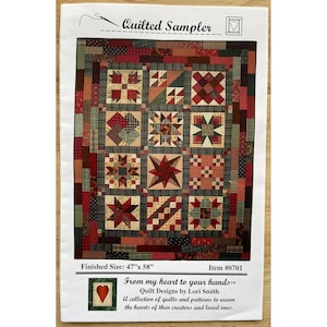 以下が含まれることがあります： 「Quilted Sampler」というタイトルのプリントキルトパターン。赤、緑、茶色のパッチワークデザイン。完成サイズは119 cm x 147 cmです。「From my heart to your hands」というテキストも表示されています。