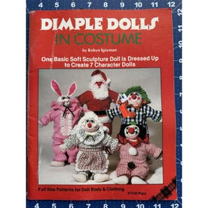 Puede incluir: Una portada de libro roja con el título "DIMPLE DOLLS IN COSTUME" e imágenes de muñecas de escultura blanda vestidas de conejo, Papá Noel y payasos. El libro es de Robyn Spizman e incluye patrones de tamaño completo.