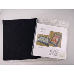 Puede incluir: Un cuadrado de tela negra junto a una bolsa de plástico transparente que contiene un papel impreso. El papel presenta el texto "Happy Grouchy" e ilustraciones de gatos. El papel también incluye el texto "CAT-itude".