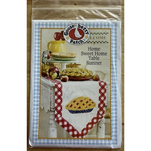 以下が含まれることがあります： 「Home Sweet Home」の文字とパイのデザインが施された、パッケージ入りのテーブルランナー。赤と白のチェック柄の縁取りと、青と白のギンガムチェックの背景があります。Gooseberry Patchのロゴが見えます。
