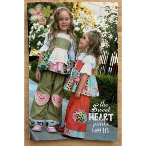 May include: Two young girls wearing matching outfits. One girl wears olive green pants with heart-shaped patches and a floral top. The other girl wears a red skirt and a floral top. The outfits are from the "Sweet Heart pants NO. 16" collection.