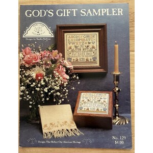 Puede incluir: Un libro de patrones de bordado de punto de cruz vintage titulado "God's Gift Sampler". La portada presenta un sampler enmarcado, una caja de madera con un diseño de sampler, una vela en un portavelas de latón y un ramo de flores rosas. El texto incluye "Designs That Reflect Our American Heritage."