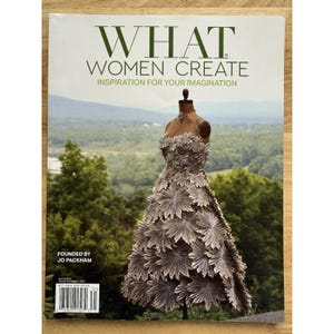 May include: The cover of "WHAT WOMEN CREATE" magazine features a dress made of folded paper on a mannequin. The magazine's title is in green, with the tagline "INSPIRATION FOR YOUR IMAGINATION." The background shows a scenic view of trees and a mountain range.