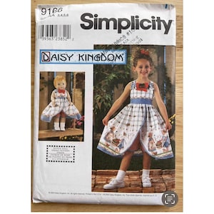 Puede incluir: Patrón de costura Simplicity con un diseño de Daisy Kingdom. El patrón incluye un vestido para una niña y una muñeca, ambos con un estampado de cuadros y detalles florales. La portada del patrón muestra a una niña usando el vestido.