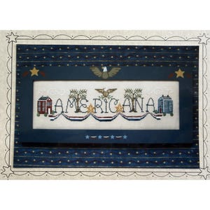 Peut inclure: Un cadre contenant une broderie au point de croix sur tissu crème avec le mot AMERICANA en noir, entouré de deux maisons, deux arbres et deux aigles. Le motif inclut des guirlandes rouge, blanc et bleu, des étoiles dorées et un cadre bleu.