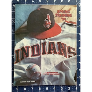 May include: A printed publication titled Spring Training '94 featuring a black and red baseball cap with a logo, a white baseball jersey with the word INDIANS in red and black lettering, and a baseball, measuring approximately 12 inches in height.