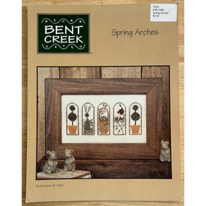 May include: A cross stitch pattern booklet titled Spring Arches by Bent Creek. The cover features a framed embroidery design with five arched panels depicting topiaries, a rabbit, a basket, and a bird, alongside two small rabbit figurines.