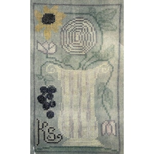 May include: A cross-stitch embroidery on light blue fabric featuring a central white vase, a yellow sunflower, a circular white floral motif, dark blue grapes, and a small white flower. The initials KS are stitched in black in the bottom left corner.