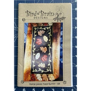 Puede incluir: Un patrón de manualidades titulado "Falling Leaves Table Runner - 169" de Bird Brain Designs. La portada presenta un diseño de camino de mesa con coloridas hojas de otoño y vides sobre un fondo oscuro.