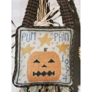 May include: A square fabric ornament featuring a cross-stitched orange jack-o'-lantern with black eyes and a jagged mouth. The design includes three yellow stars, the text PUMPKIN, and the year 1994 in blue, finished with a black plaid border.