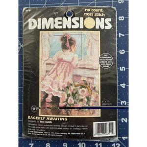 Peut inclure: Un kit de point de croix Dimensions intitulé "Eagerly Awaiting". Le kit présente un motif d'une jeune fille en robe rose regardant par une fenêtre un panier de fleurs. Le paquet mesure 13cm x 18cm.