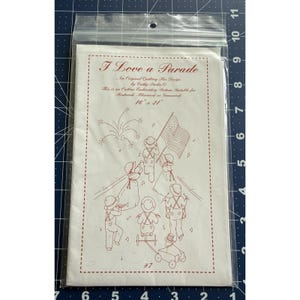 May include: A clear plastic bag containing a white paper with a red outline embroidery pattern titled "I Love a Parade." The pattern features marching figures, an American flag, and fireworks. The paper measures 16 x 21 inches.
