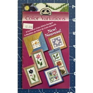 May include: A DMC Color Variations cross-stitch pattern booklet featuring six exclusive designs. The magenta cover displays images of finished embroidery projects including flowers, hearts, and nautical themes with the text New! Nouveau! and $2.00.
