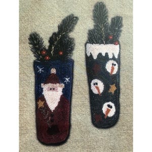 May include: Two vertical punch needle wall hangings featuring holiday themes. The left piece depicts a Santa Claus figure in a maroon coat against a dark blue background. The right piece shows three snowmen on a black background with white trim.