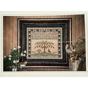May include: A framed cross-stitch sampler with a brown wooden frame. The sampler features a tree with red fruit, a bird, and text that reads "With careful hands our work complete..." The year 2003 is also visible. Decorative elements include a small bird figurine and greenery.