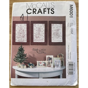Op de afbeelding: McCall's Crafts patroon M6001 met drie ingelijste kerstontwerpen. Het patroon bevat een kerstboom, de kerstman en een sneeuwpop. Extra kerstversieringen worden getoond op een witte tafel.
