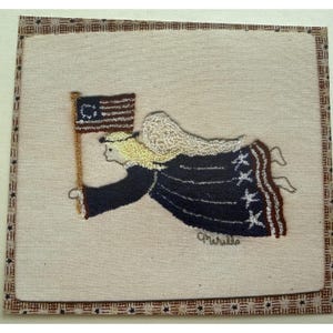 Pode incluir: Uma tapeçaria quadrada de tecido com bordado de agulha mágica representando um anjo de cabelo loiro e asas brancas, vestindo um vestido azul-escuro com estrelas brancas e segurando uma bandeira de Betsy Ross. Borda com padrão de estrelas.
