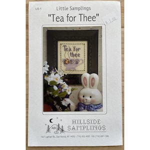 May include: A cross-stitch pattern leaflet titled Little Samplings Tea for Thee. The cover features a framed cross-stitch of a teacup with the text Tea for Thee, a small white ceramic bunny figurine with a blue bow, and a bouquet of white flowers.