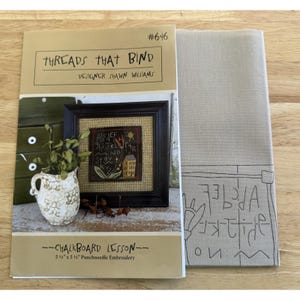 Może przedstawiać: Zestaw rzemieślniczy z tytułem "THREADS THAT BIND" i nazwiskiem projektanta Shawn Williams. Zestaw zawiera oprawiony haft igłowy z motywem domu i alfabetu oraz beżową tkaninę z wyszywaną obwódką. Zestaw ma wymiary 14 cm x 14 cm.