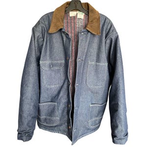 May include: A blue denim jacket with a brown corduroy collar. The jacket has four front pockets, white stitching, and a button-up closure. The interior lining features a red and white striped pattern. The jacket is hanging on a black hanger.