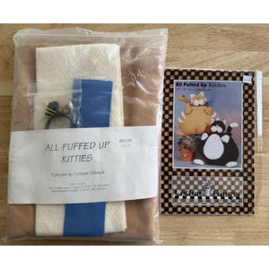 May include: A craft kit for making stuffed kitties. The kit includes fabric pieces in beige, brown, and blue, along with embellishments. The packaging features the text "ALL PUFFED UP KITTIES" and a pattern by Cotton Ginny's.