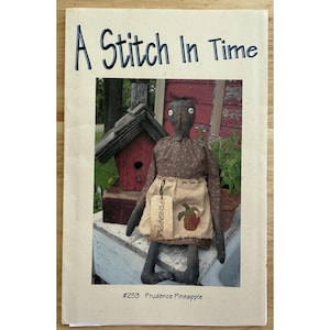Puede incluir: Portada del patrón A Stitch In Time con una muñeca de arte popular llamada Prudence Pineapple. La muñeca viste un vestido con estampado floral y un delantal con diseño de piña. El fondo incluye una casita para pájaros y vegetación.