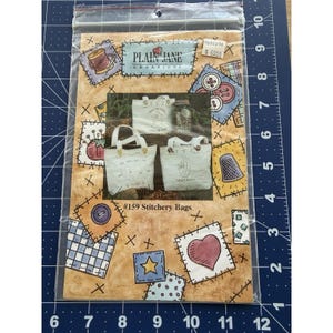 May include: A package featuring a sewing pattern for #159 Stitchery Bags. The design includes images of tote bags, buttons, spools of thread, and a heart. The words "PLAIN JANE CREATIONS" are visible. The background is tan with patchwork-style accents.