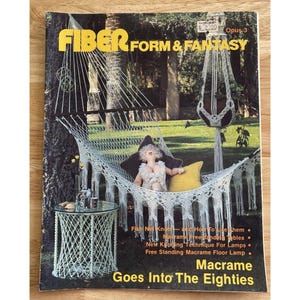 May include: The cover of a vintage magazine titled "Fiber Form & Fantasy" features a macrame hammock with a doll and yellow pillow. The magazine cover also shows a macrame plant hanger and a small table. The text "Macrame Goes Into The Eighties" is visible.