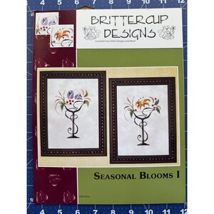 以下が含まれることがあります： Brittercup Designsの「Seasonal Blooms I」というタイトルのクロスステッチパターンパッケージ。パッケージには、装飾的なゴブレットに花のアレンジメントが施された2つの額入りクロスステッチデザインが掲載されています。カラーパレットには、緑、紫、オレンジ、白の色合いが含まれています。