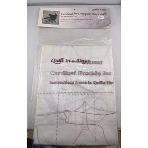 Puede incluir: Un paquete de plástico transparente que contiene un patrón de acolchado. El patrón es para un diseño de cardenal, con el texto "Quilt in a Day" y "Cardinal Fusible for" visibles. El paquete también incluye el texto "Cardinals for Lodgepole Pine Fusible".