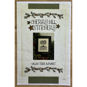 May include: A cream-colored cross-stitch pattern featuring the text "Cherry Hill Stitchery" and "Willow Tree Alphabet." The design includes a framed tree image and decorative star and leaf accents in brown and green.