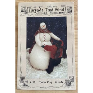 May include: A craft pattern titled "Threads That Bind" features a snowman and a child playing in the snow. The snowman is white with a plaid scarf and the child is wearing a red sweater. The pattern is 11 inches tall.