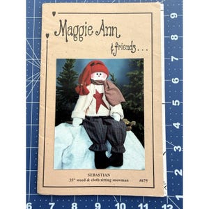 Könnte beinhalten: Ein Schnittmusterumschlag mit einem sitzenden Schneemann namens Sebastian. Der Schneemann trägt einen roten Hut, einen gemusterten Schal und einen Pullover mit einem roten Stern. Der Text "Maggie Ann & friends..." steht oben. Der Schneemann ist 89 cm groß.