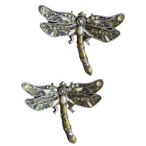 Peut inclure: Deux broches libellules. Chaque broche présente un design détaillé avec des accents verts, blancs et argentés. Les ailes sont ornées de petits cristaux étincelants. Le corps de la libellule est un mélange de vert et d'argent, avec une perle au centre.